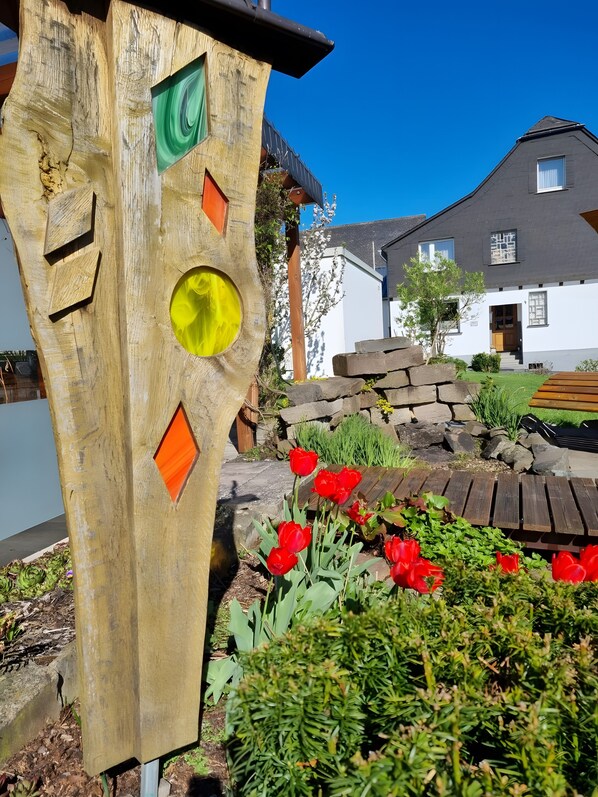 A wooden sculpture with geometric shapes, a garden with red flowers, and a white building with a dark roof in the background.