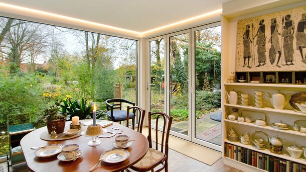Un comedor con una mesa dispuesta para dos, una estantería con adornos y vistas a un jardín a través de grandes ventanales.