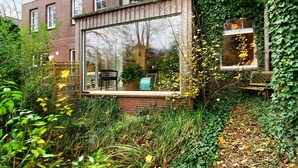 Una casa moderna con un amplio ventanal, un muro de ladrillo y un jardín con diversas plantas.