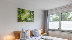 Une chambre à coucher comprenant un lit, deux tables de chevet, une lampe de chevet, une horloge murale, une fenêtre avec stores et une décoration murale sur le thème de la forêt.