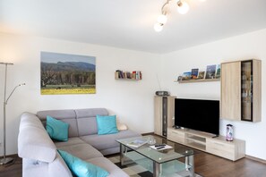 Ein modernes Wohnzimmer mit einem Ecksofa, einem Glastisch, einem Flachbildfernseher und einem Bücherregal mit Reiseführern.