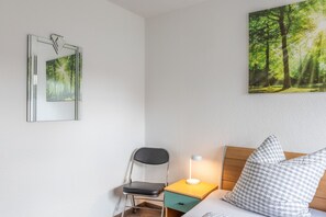 Ein Schlafzimmer mit einem Bett, einem Nachttisch mit Lampe, einem Sessel, einer kleinen Schublade und zwei an der Wand hängenden Bildern.