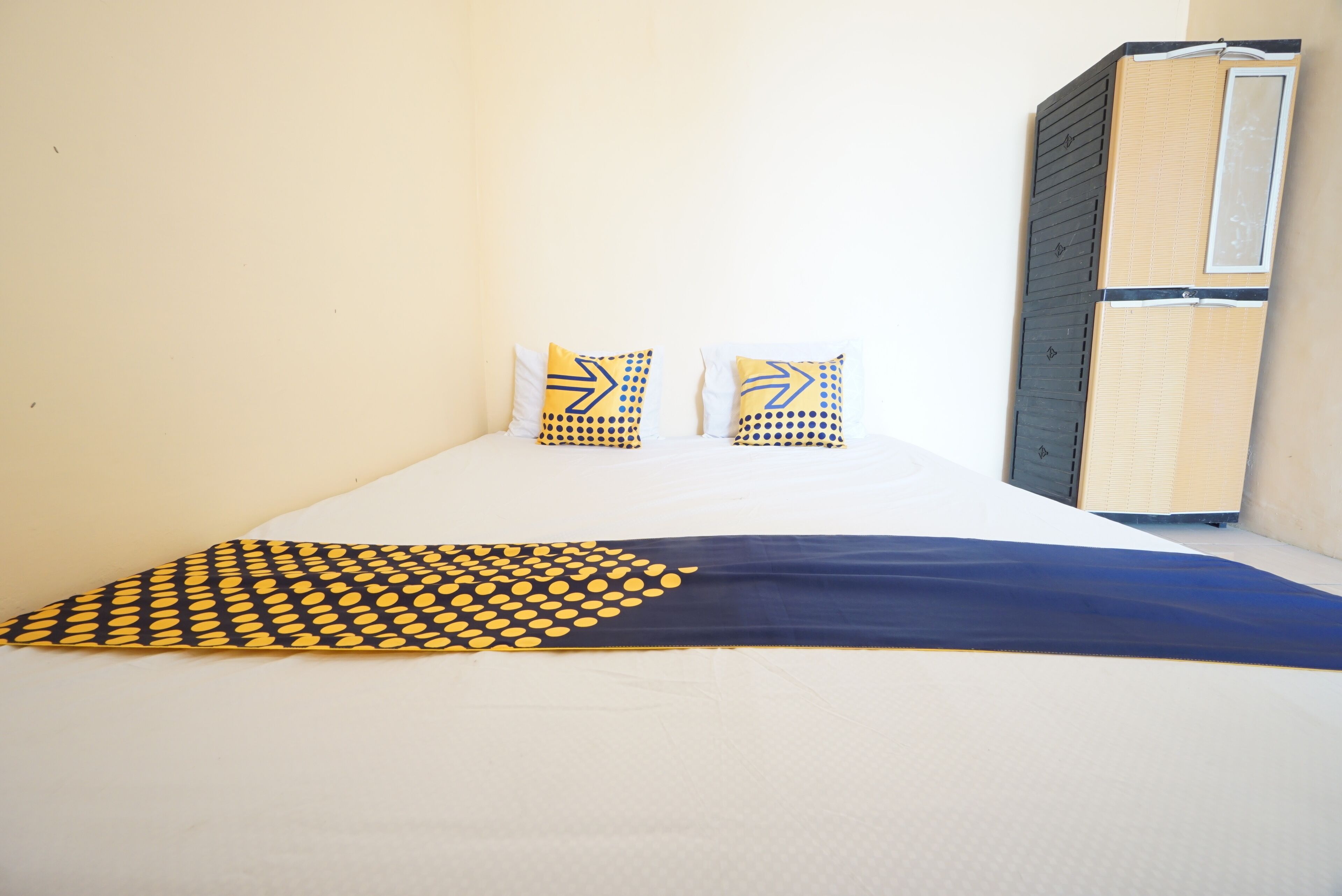 A bed with a patterned blanket, two pillows with geometric designs, and a wardrobe against a plain wall.