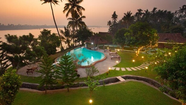 A pool area with a curved edge, surrounded by a well-manicured lawn, palm trees, and a view of the water body at dusk. - Ananda Cherai (Kochi)