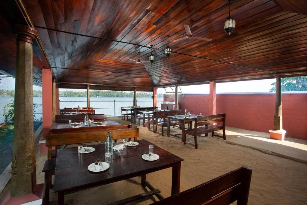 An outdoor dining area with wooden tables and chairs, a view of a body of water, and a red wall in the background.