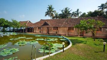 A traditional building with a tiled roof, a pond with lily pads, and a grassy area with palm trees.