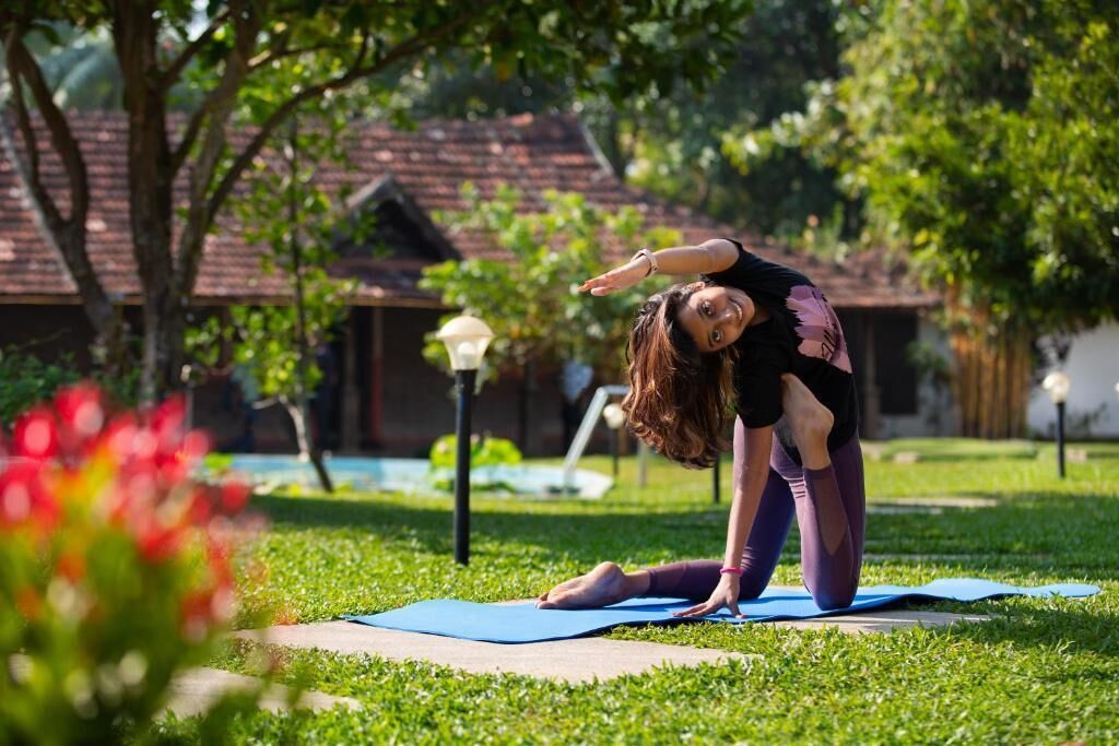 A person performing a yoga pose on a mat in a grassy outdoor area with trees and a building in the background.