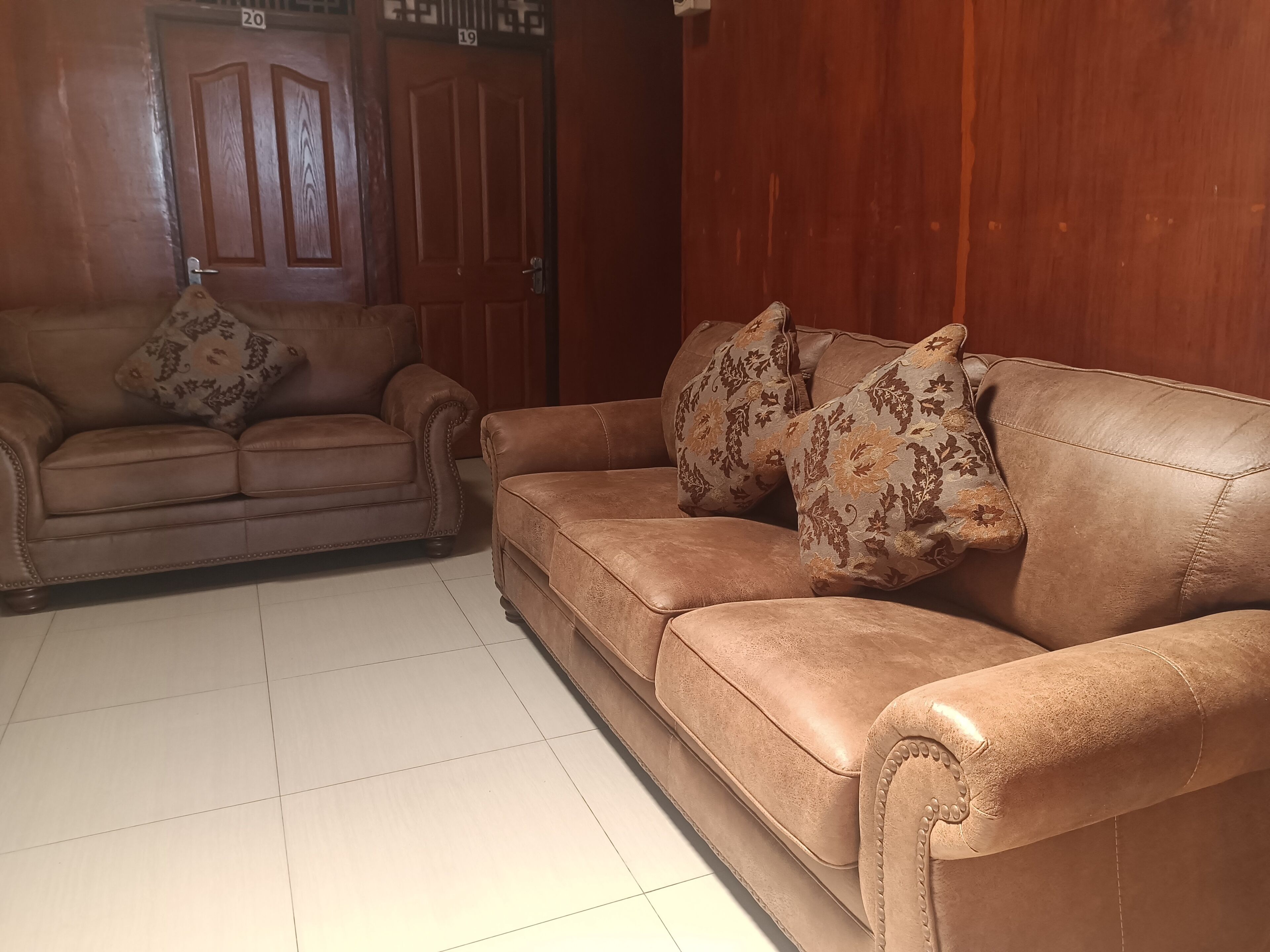 A living room with two brown leather sofas, each with patterned cushions, and a wooden door with numbers 19 and 20 above it.