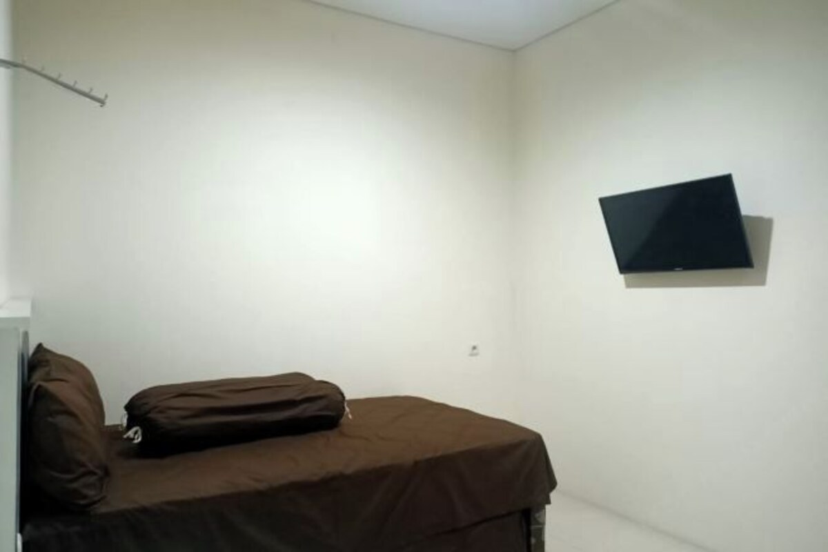 A small, single-bed room with a brown bedspread, a mounted television on the wall, and a coat rack.