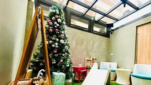 Una stanza con prato sintetico verde, uno scivolo bianco, un’altalena in legno, un albero di Natale e una finestra con vista sugli edifici.