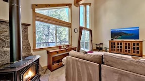 A cozy room with a stone fireplace, large windows, a sofa, and a television. - Donner Adventure Lodge: Pet Friendly, Games, Bunks (Truckee)