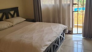 A bedroom with a bed, curtains, and a view of a garden through a window. - Dream Day Cottages (Kampala)