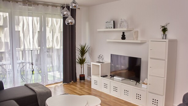Una sala de estar moderna con televisor de pantalla plana, muebles blancos, una mesa de comedor y un ventanal con cortinas translúcidas.