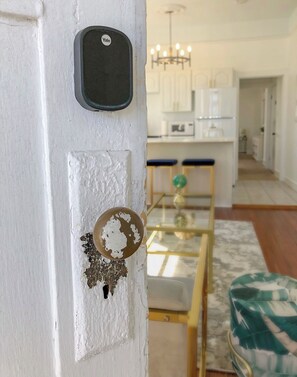 A white door with a Yale lock, a glass door handle, and a partially visible kitchen area with white cabinets and a chandelier.