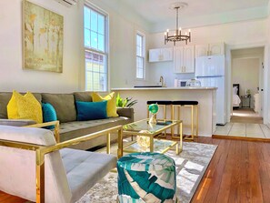 A modern living room with a sofa, coffee table, and ottoman, a kitchen with white cabinets, and a dining area with a glass table.