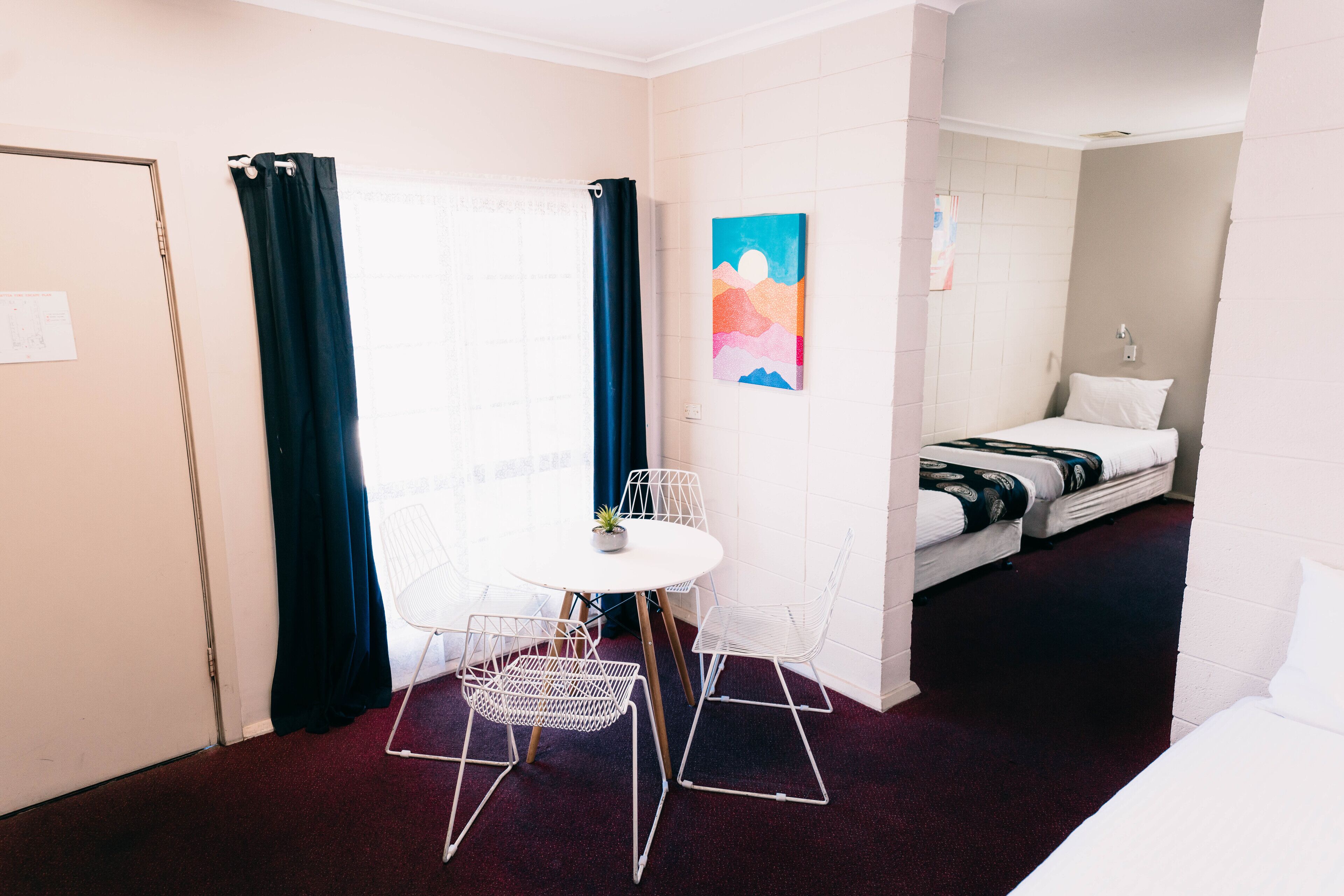 A compact room with a bed, a small table with chairs, a window with curtains, and a painting on the wall.