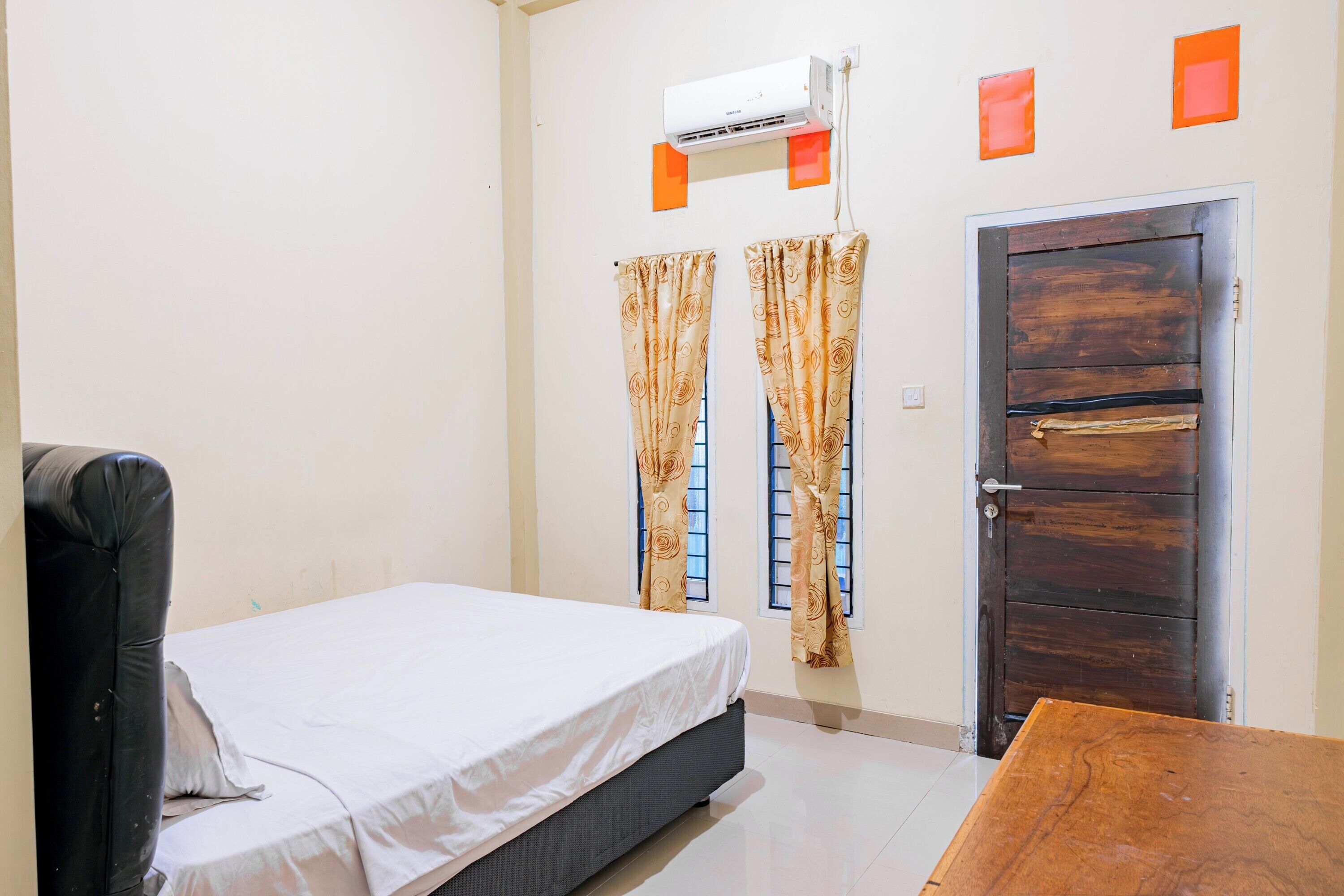 A bedroom with a bed, a wooden table, a black chair, a window with curtains, and a door.