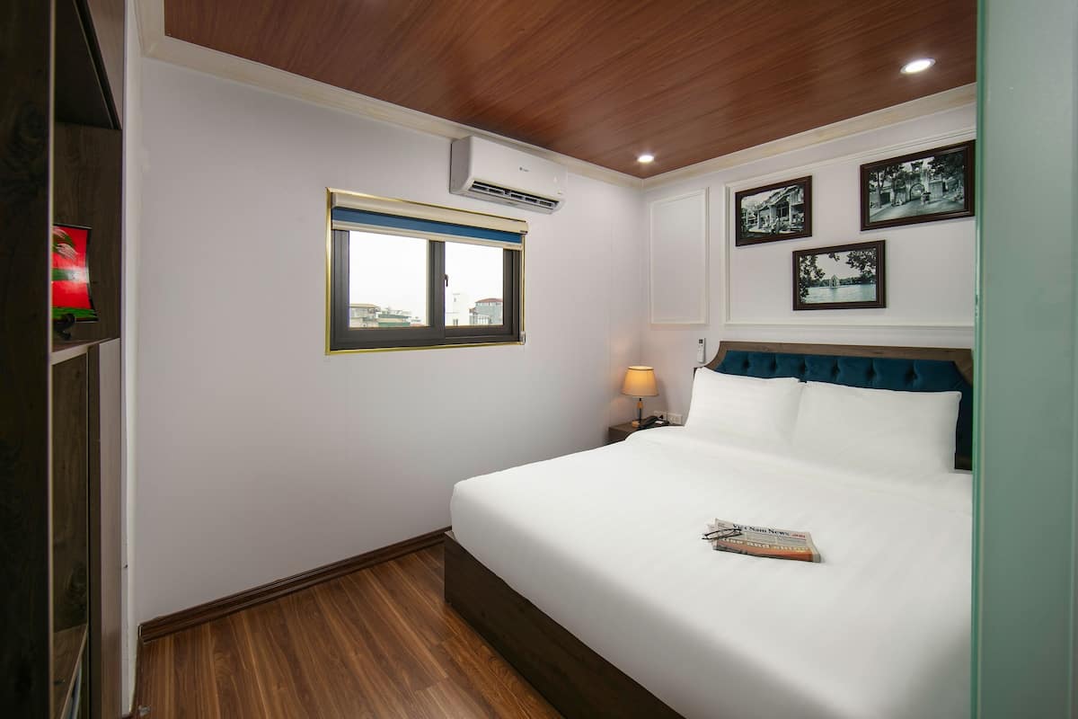 A hotel room with a wooden ceiling, a large bed with white bedding, a nightstand with a lamp, and three framed pictures on the wall.