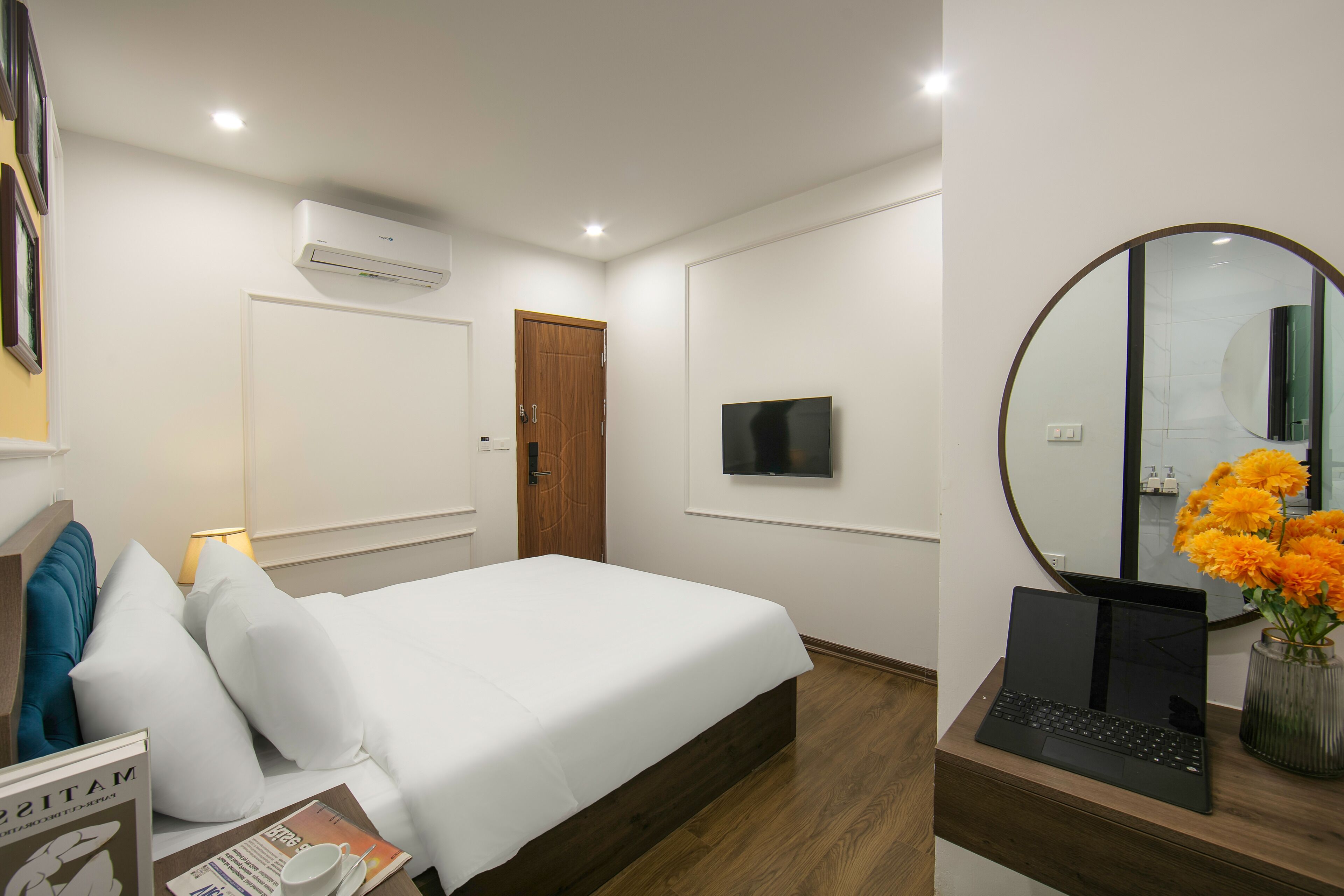 Una habitación de hotel con una cama, televisión, computadora portátil, espejo y un florero con flores.