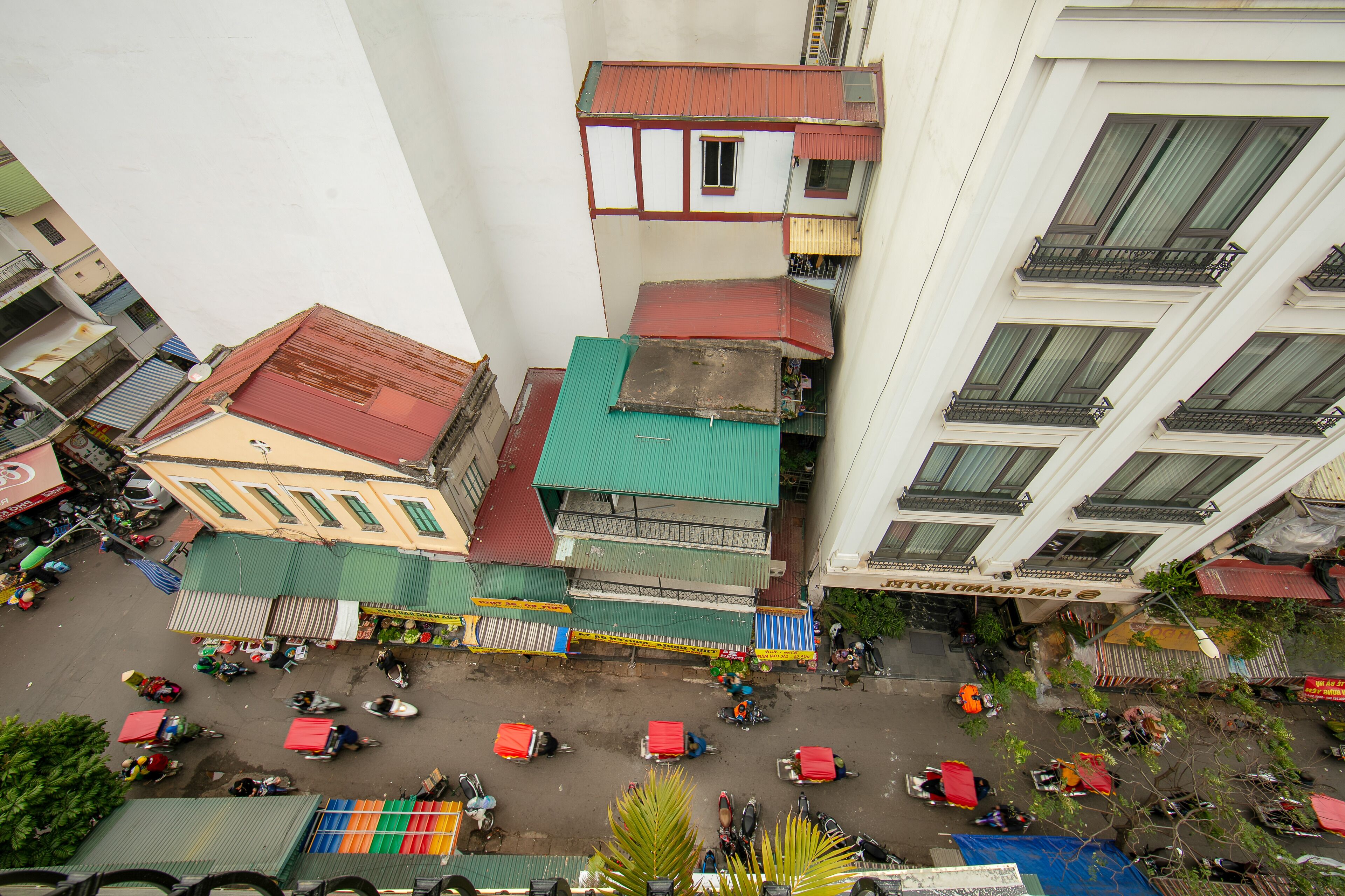 Un immeuble à plusieurs étages doté de balcons, une rue en contrebas où sont garés des véhicules, et divers commerces.