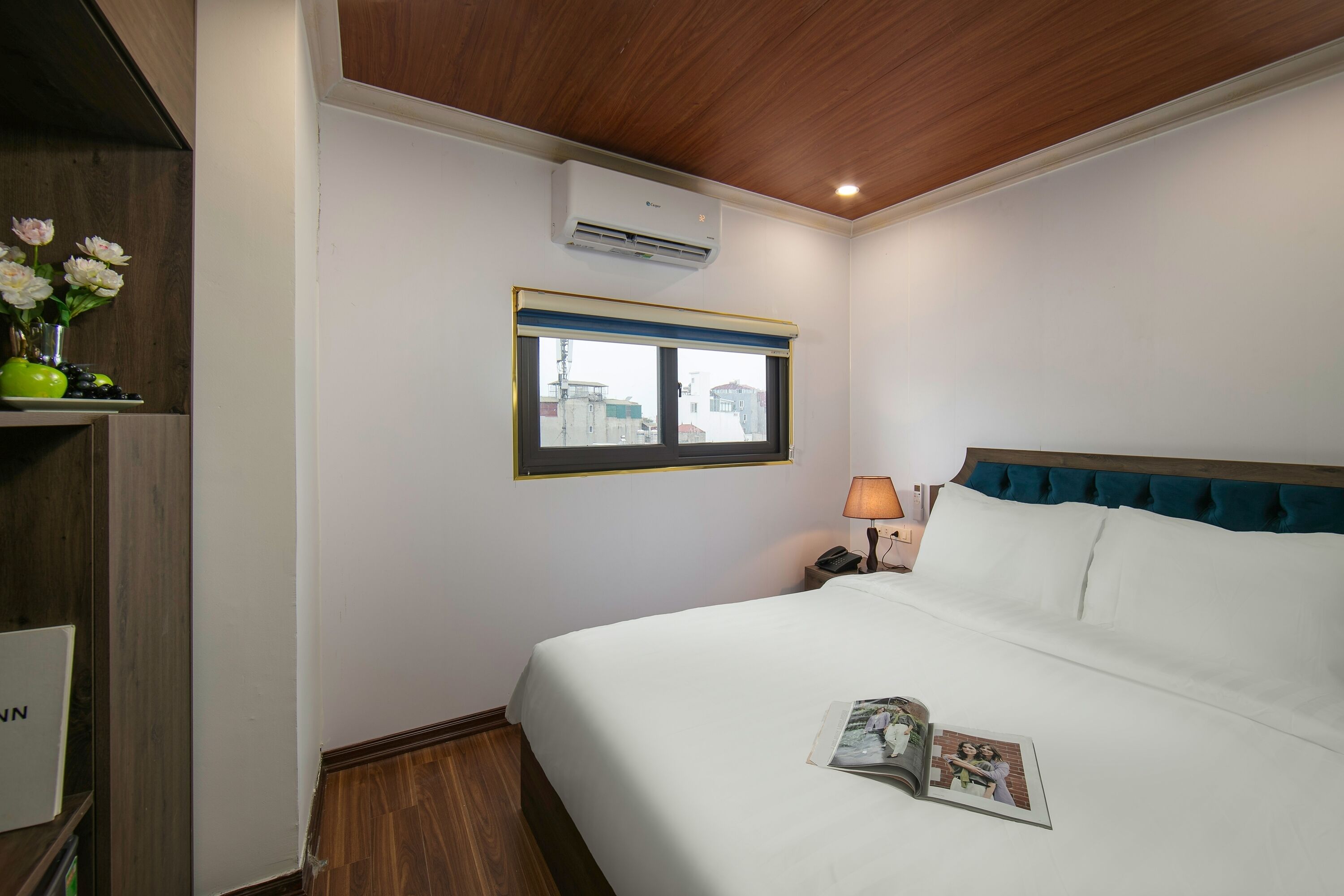A hotel room with a bed, a nightstand with a lamp, a window with a view, and an air conditioning unit.