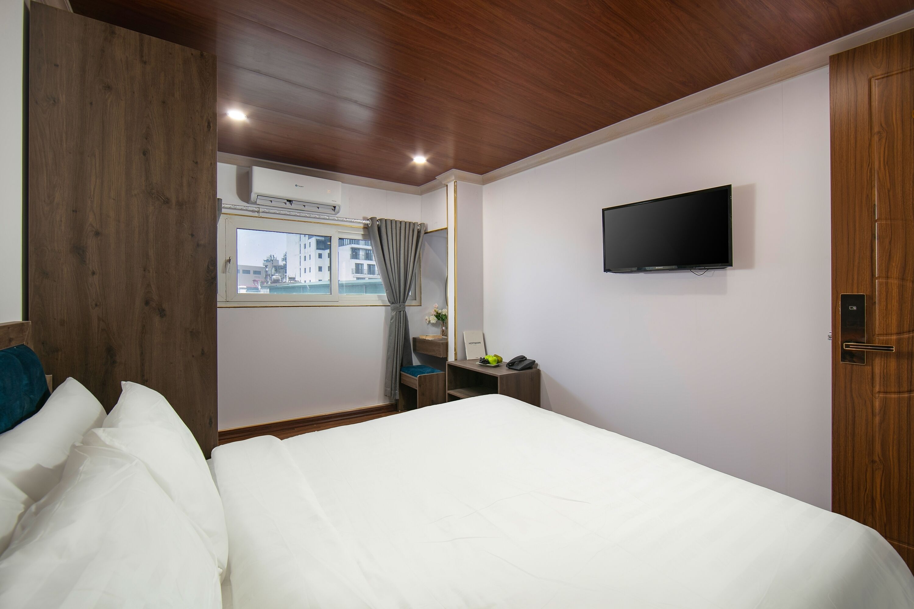 A hotel room with a wooden ceiling, a bed with white linens, a desk with a chair, a flat-screen TV mounted on the wall, and a window with curtains.