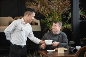 Um garçom servindo uma xícara de café a um cliente sentado em um ambiente interno moderno, com uma planta grande e uma câmera sobre a mesa.