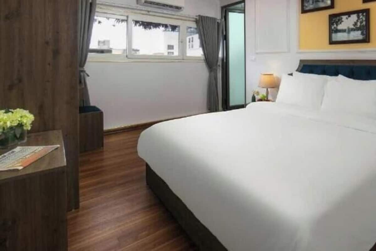 A hotel room with a large bed, wooden flooring, a small table with a vase of flowers, a nightstand with a lamp, and a window with curtains.