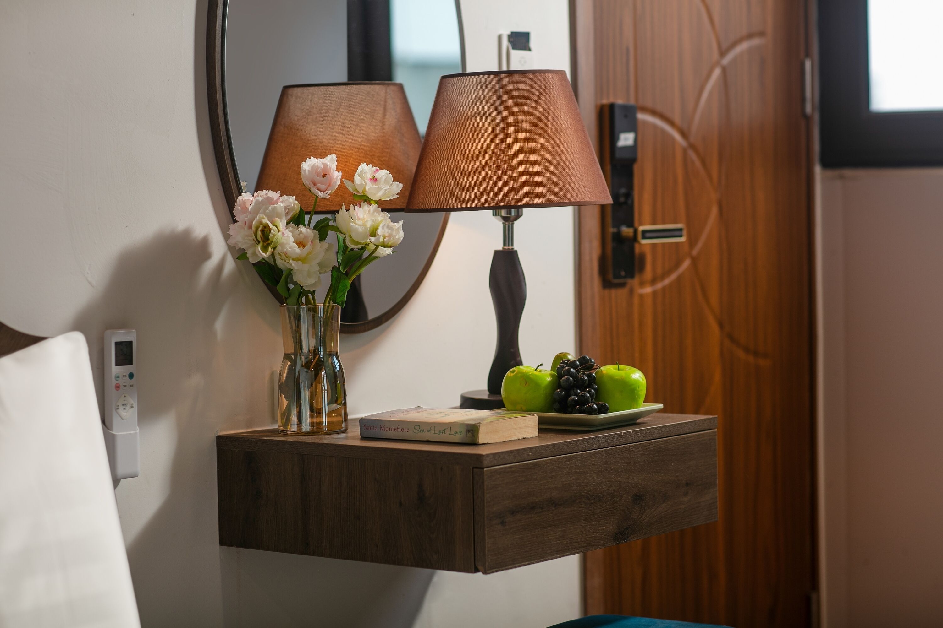 A bedside table with a book, a vase of flowers, and a plate of fruit.