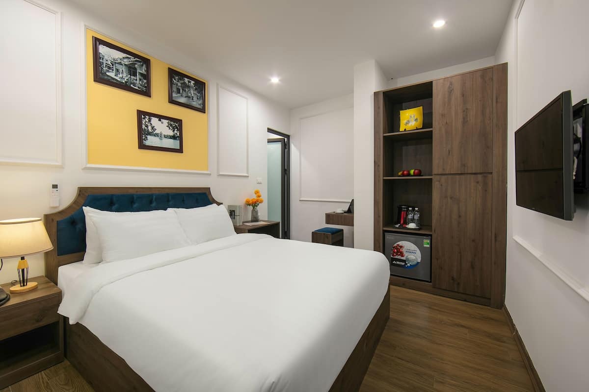 A neatly arranged hotel room with a large bed, a bedside table with a lamp, a wardrobe with shelves, and framed pictures on the wall.