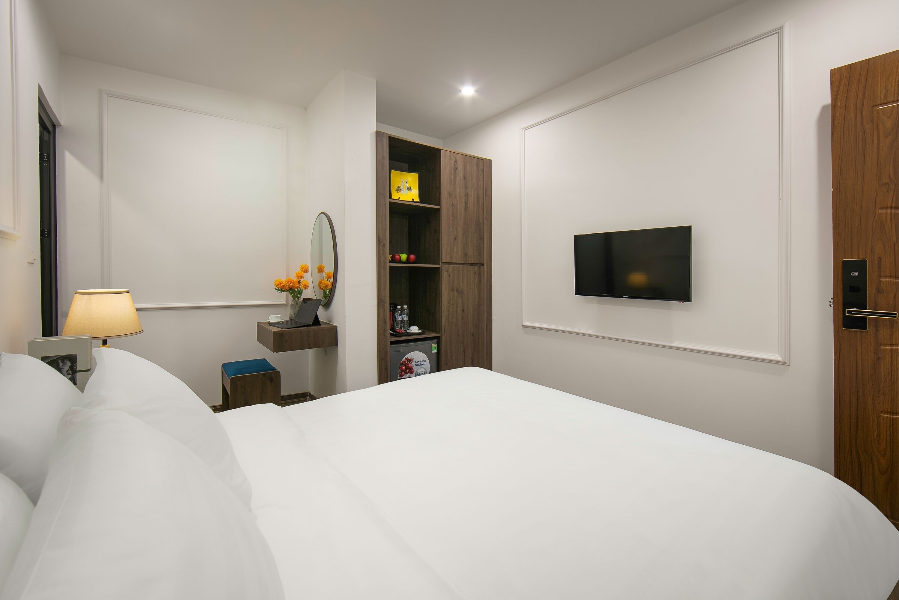 Ein Hotelzimmer mit einem großen Bett, einem an der Wand montierten Fernseher, einem Holzschrank, einem kleinen Tisch mit Lampe und einem Regal mit Dekoration.
