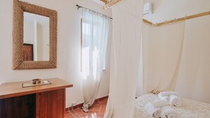 A bedroom with a wooden desk, a mirror, a bed with rolled towels, and a window with sheer curtains.