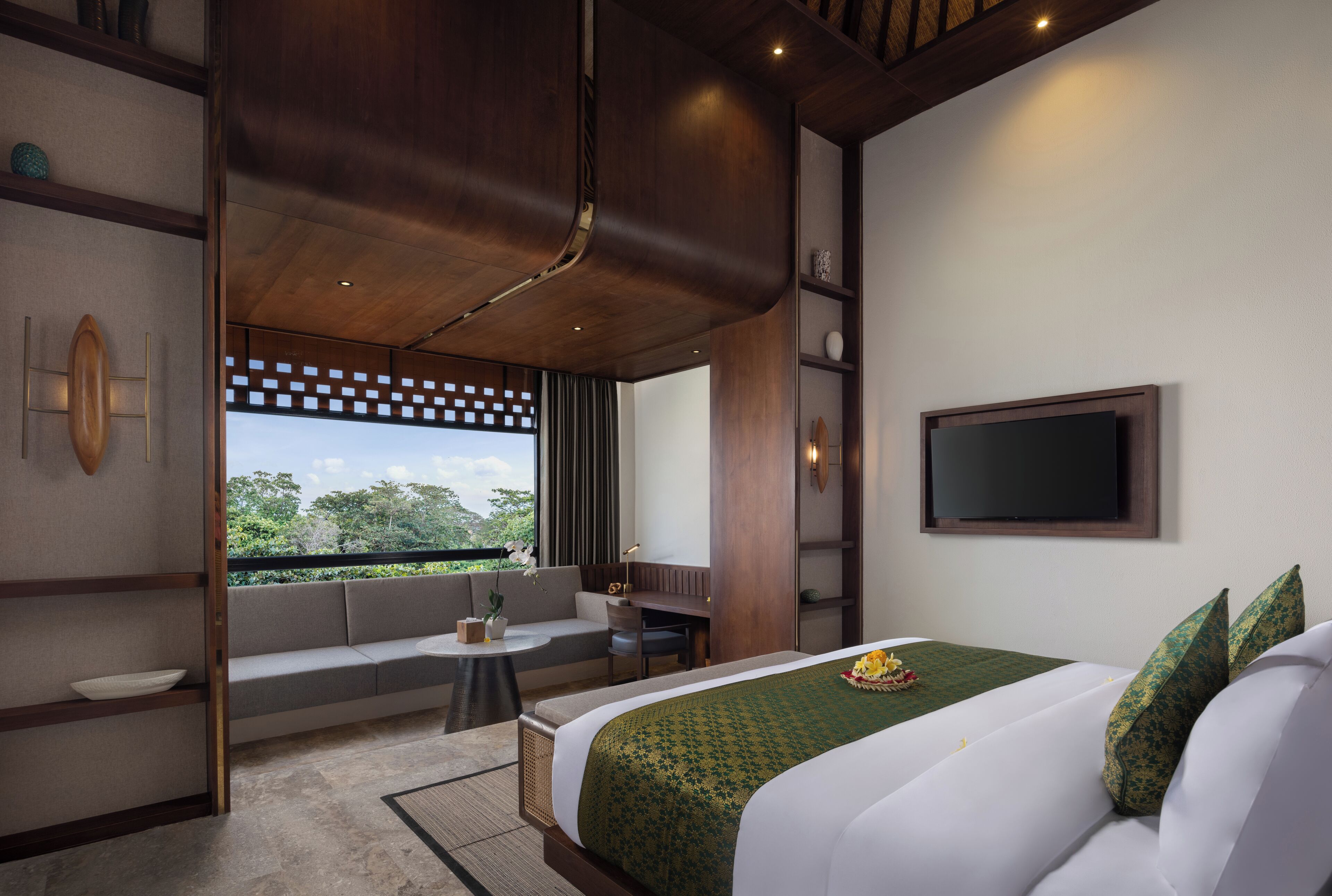 A modern hotel room with a large bed, a seating area with a sofa and a small table, a TV mounted on the wall, and a window with a view of greenery.