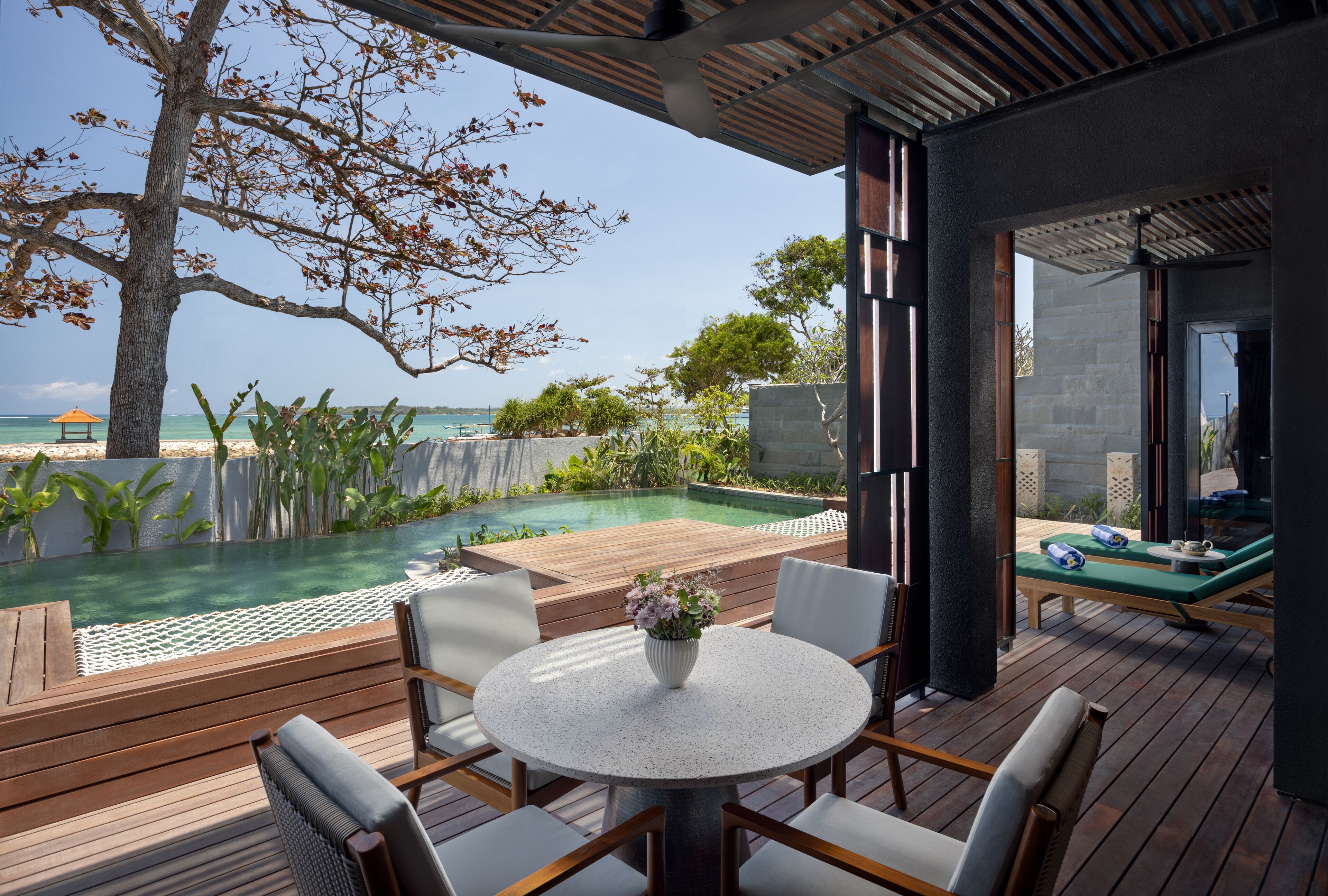 Una terraza de madera con una mesa redonda y sillas, con vista a una piscina y al océano.
