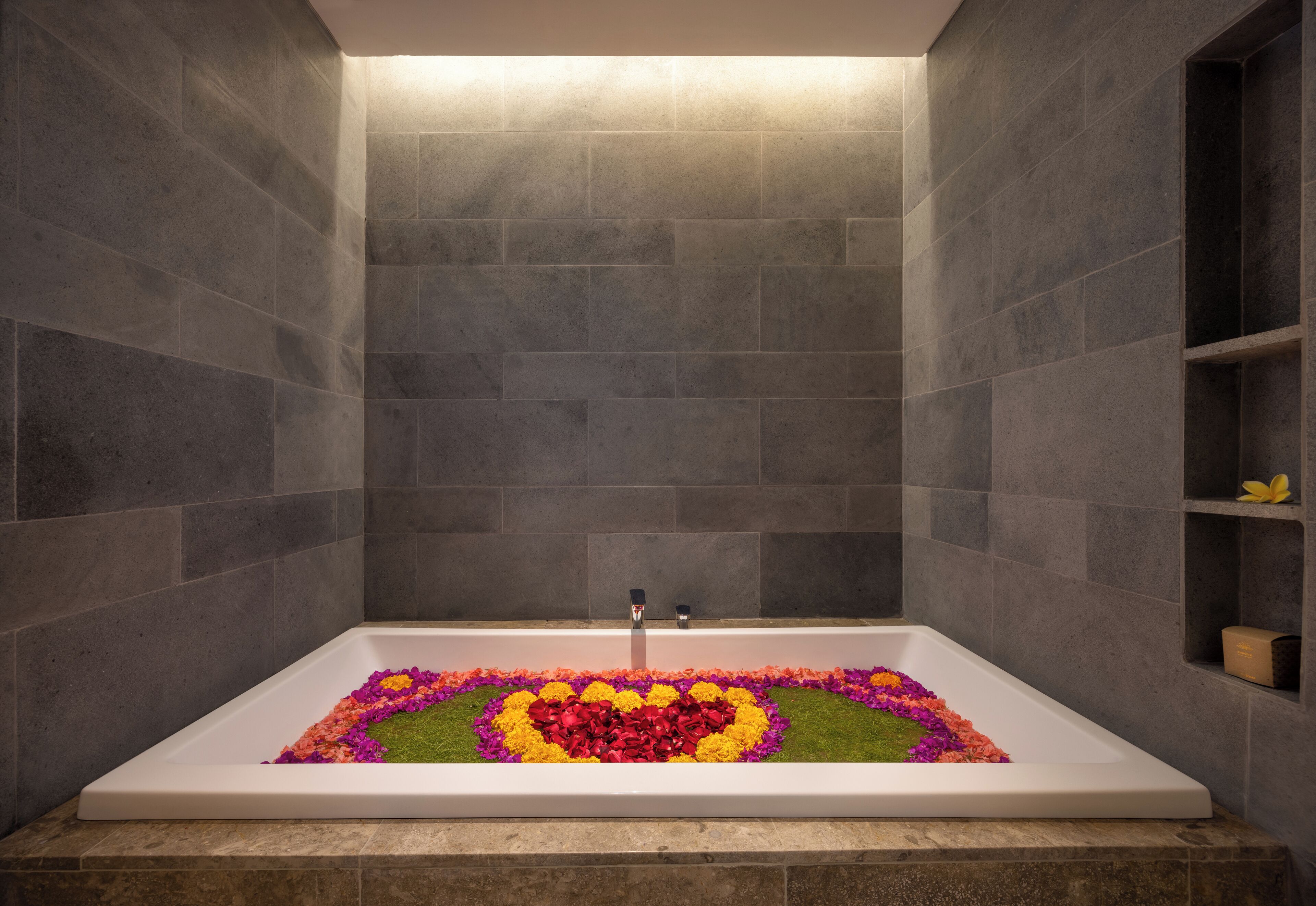 A rectangular bathtub with a heart-shaped floral arrangement on the floor, surrounded by grey stone walls.