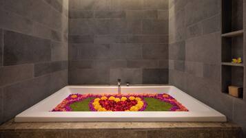 Una bañera rectangular con un arreglo floral en forma de corazón sobre el piso, rodeada de paredes de piedra gris.