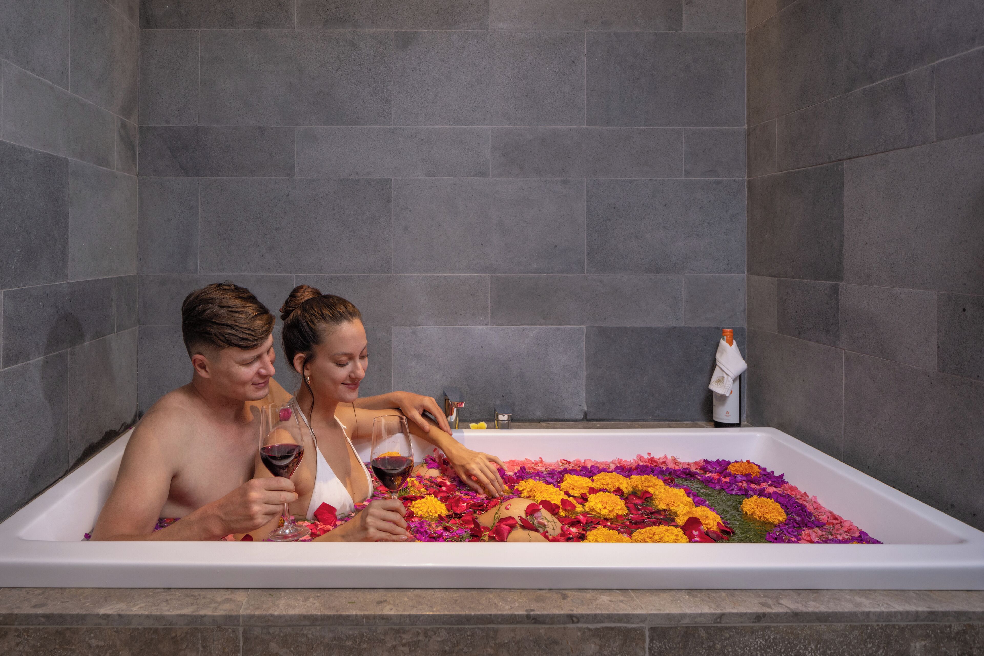 A bathtub filled with flowers and a person holding a glass.