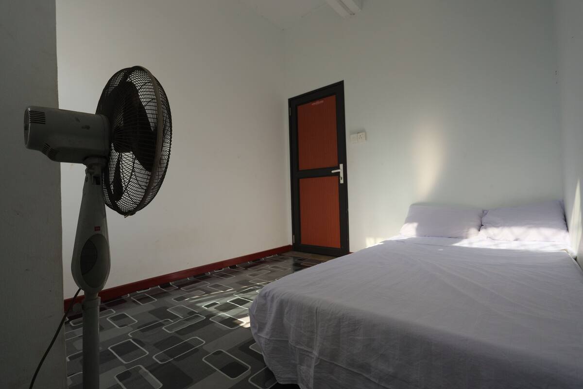 A room with a bed, a standing fan, and a door.
