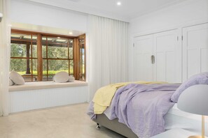 Ein Schlafzimmer mit einem großen Fenster, einem Bett mit gelb-lila Decke und weißen Wänden.