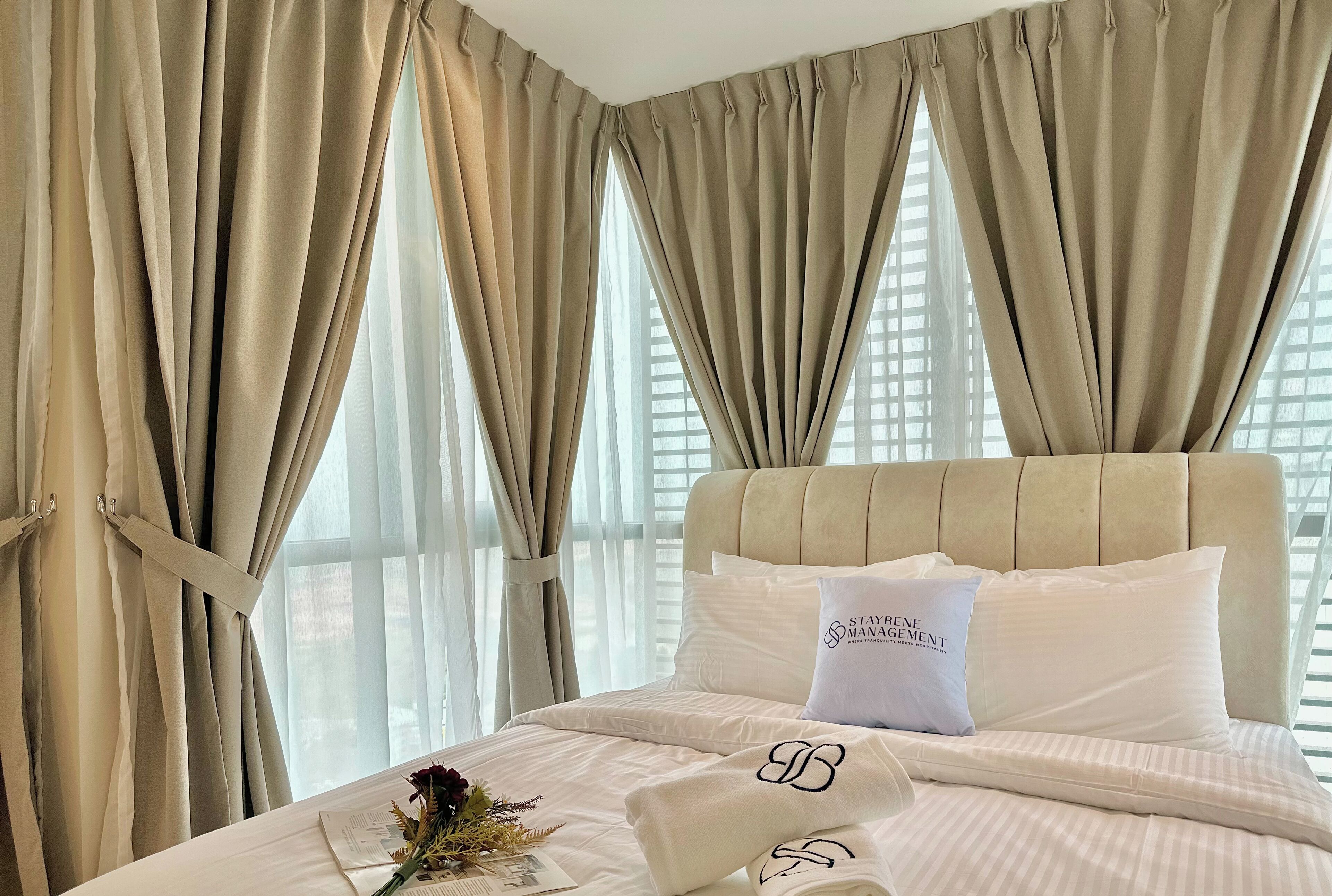 A hotel room with a large bed, beige curtains, and a view of the city through the windows.
