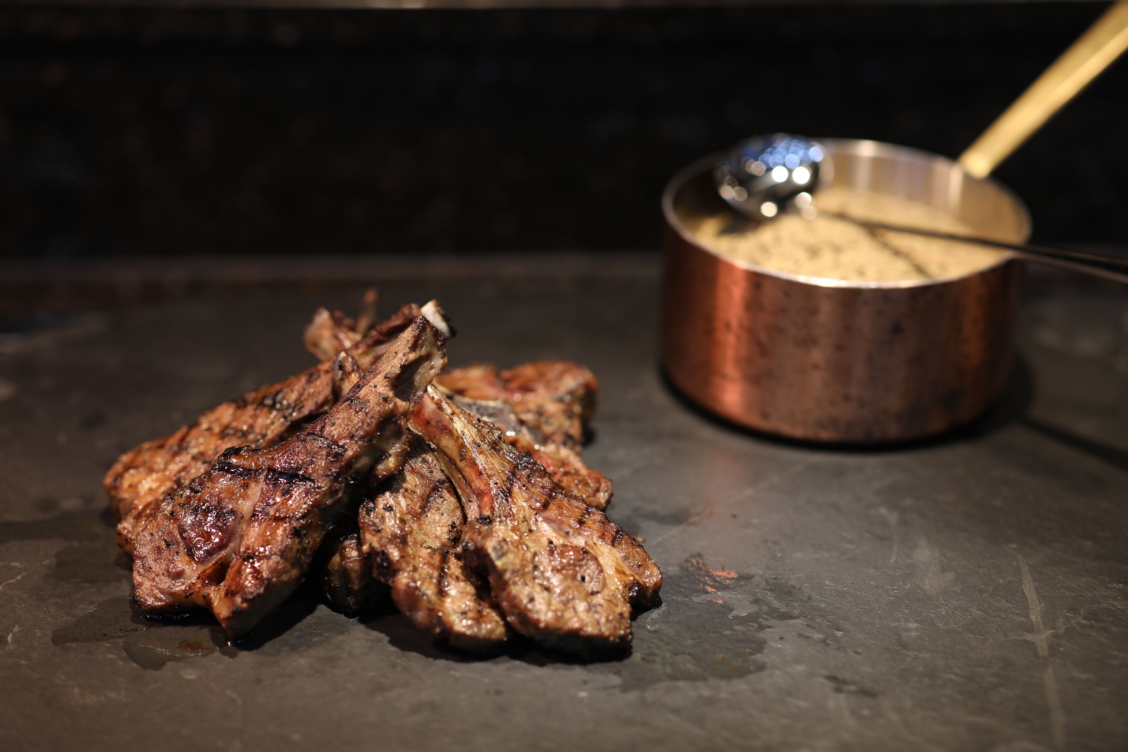 Grilled meat and a pot with a ladle on a dark surface.