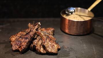 Grilled meat and a pot with a ladle on a dark surface.