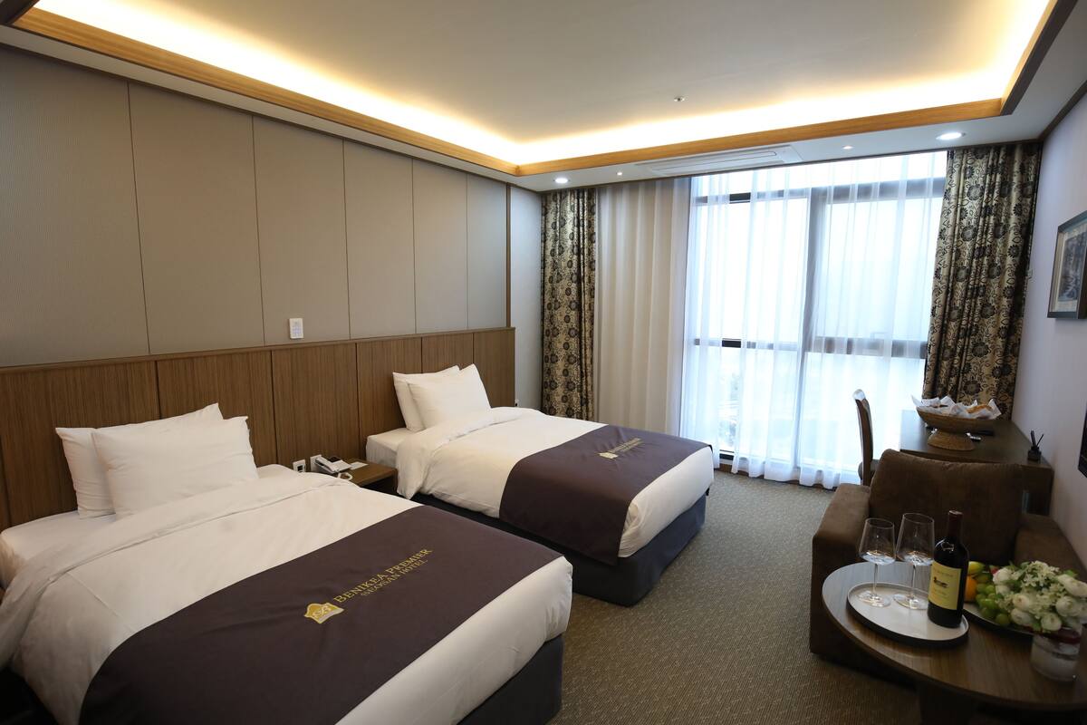 A hotel room with two beds, a desk with a vase of flowers, and a view of the outside through large windows.