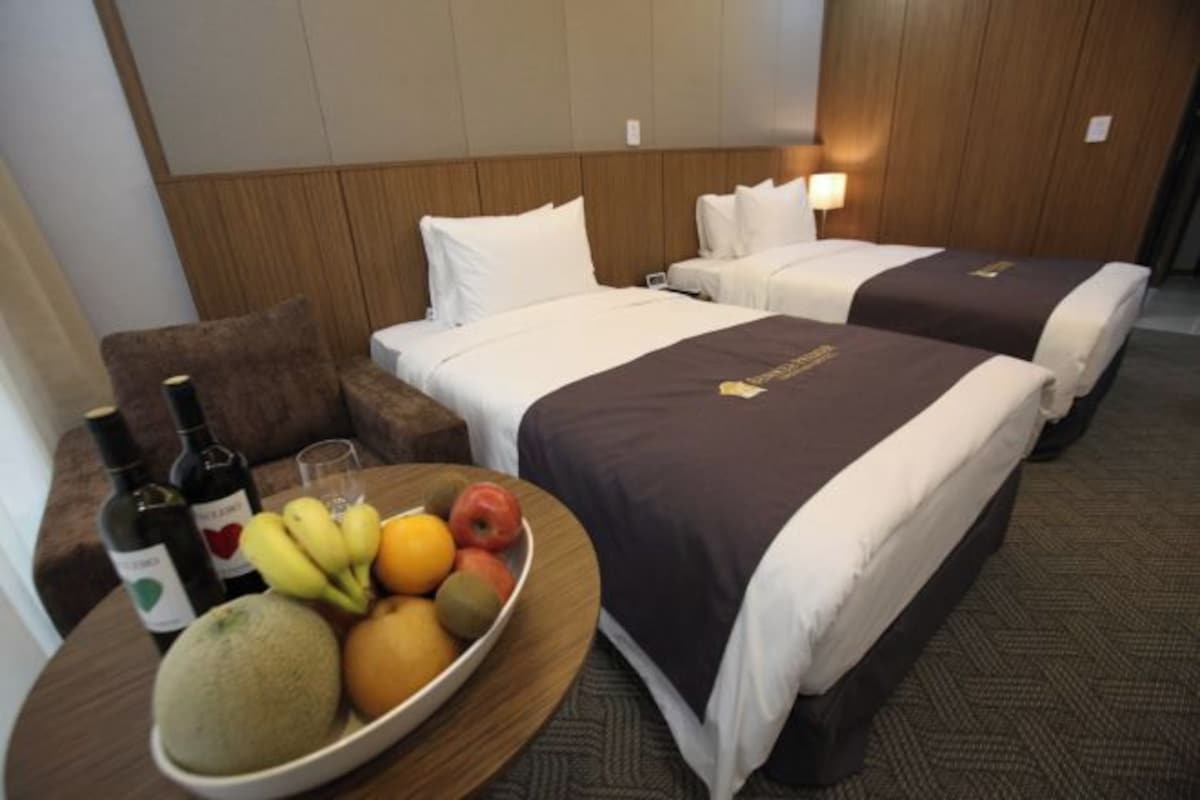 A hotel room with two beds, a dining table with fruit and wine, and a nightstand.