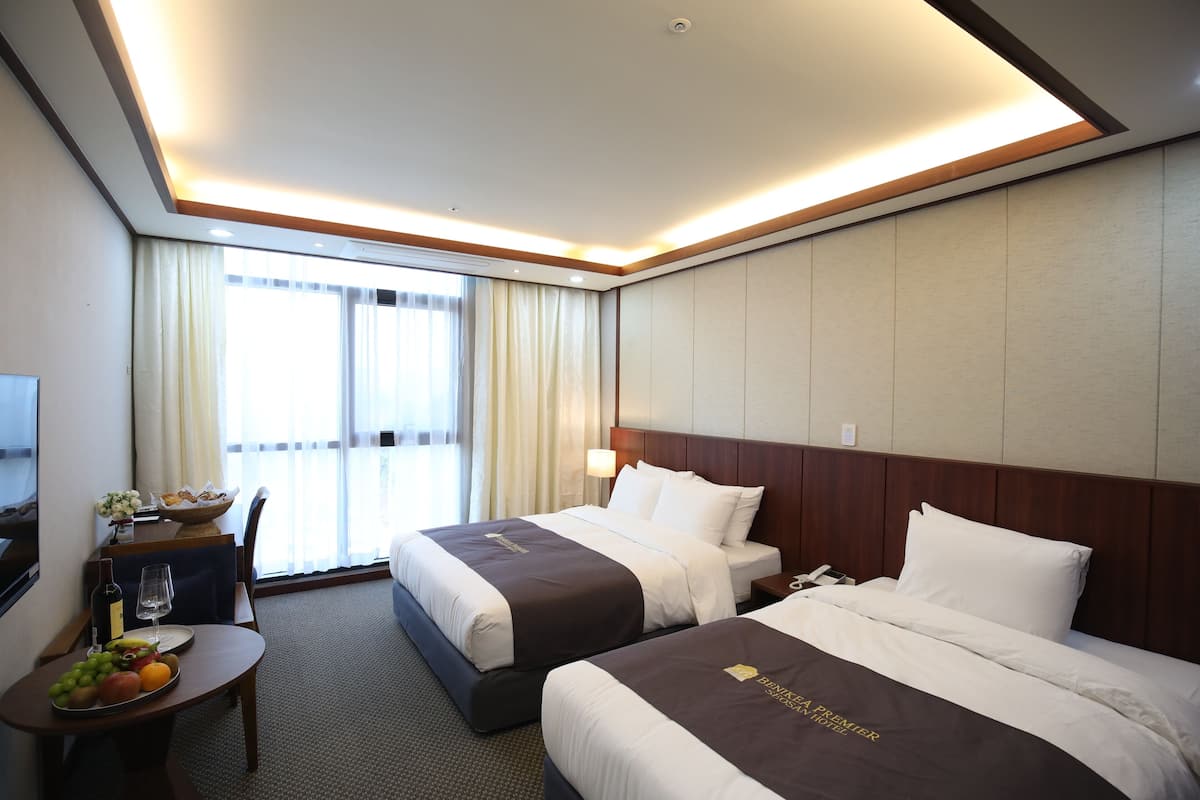 A hotel room with two beds, a desk with a fruit bowl, a bottle of wine, and a lamp.