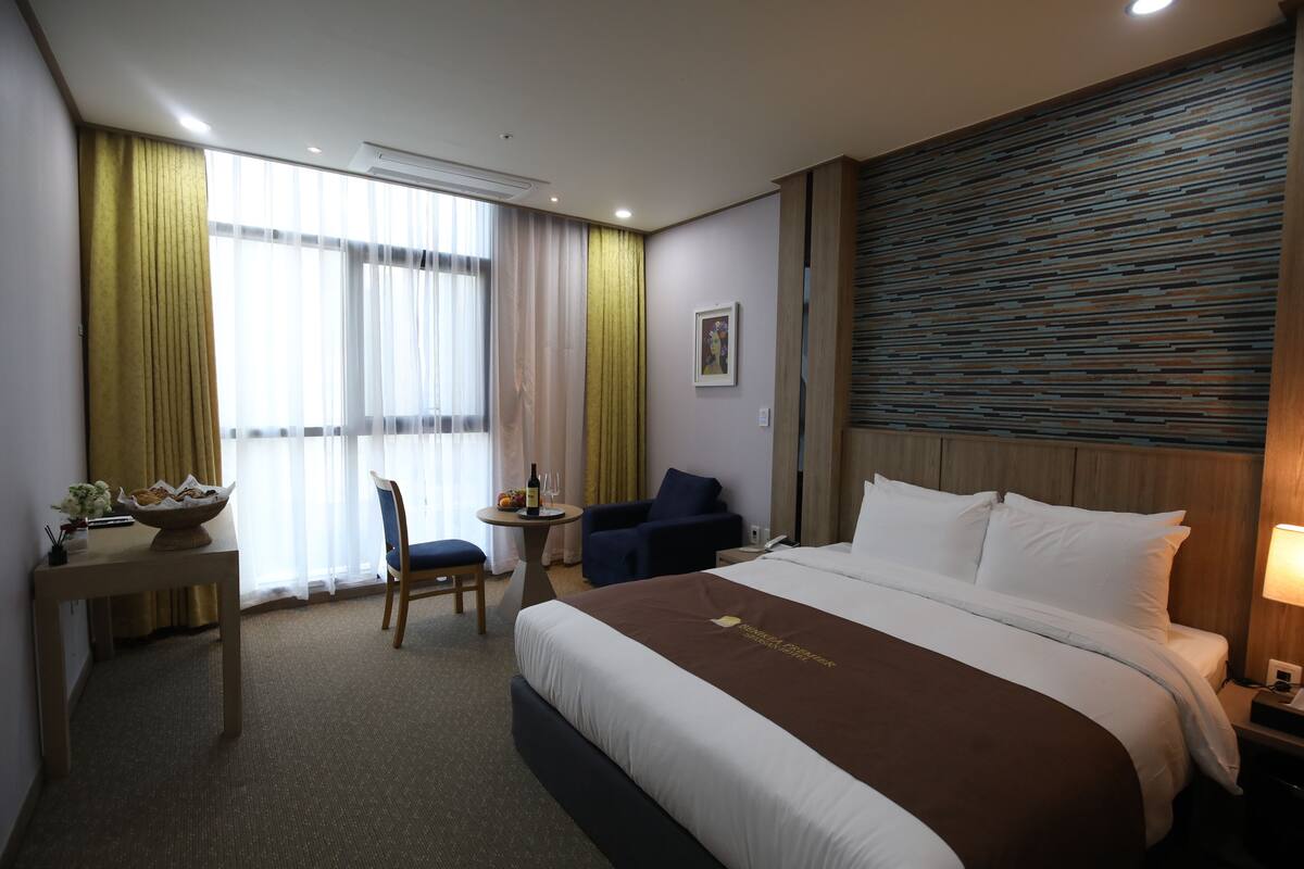 A hotel room with a large bed, a desk with a bowl of fruit, a chair, a small table with a bottle and glasses, and a window with curtains.