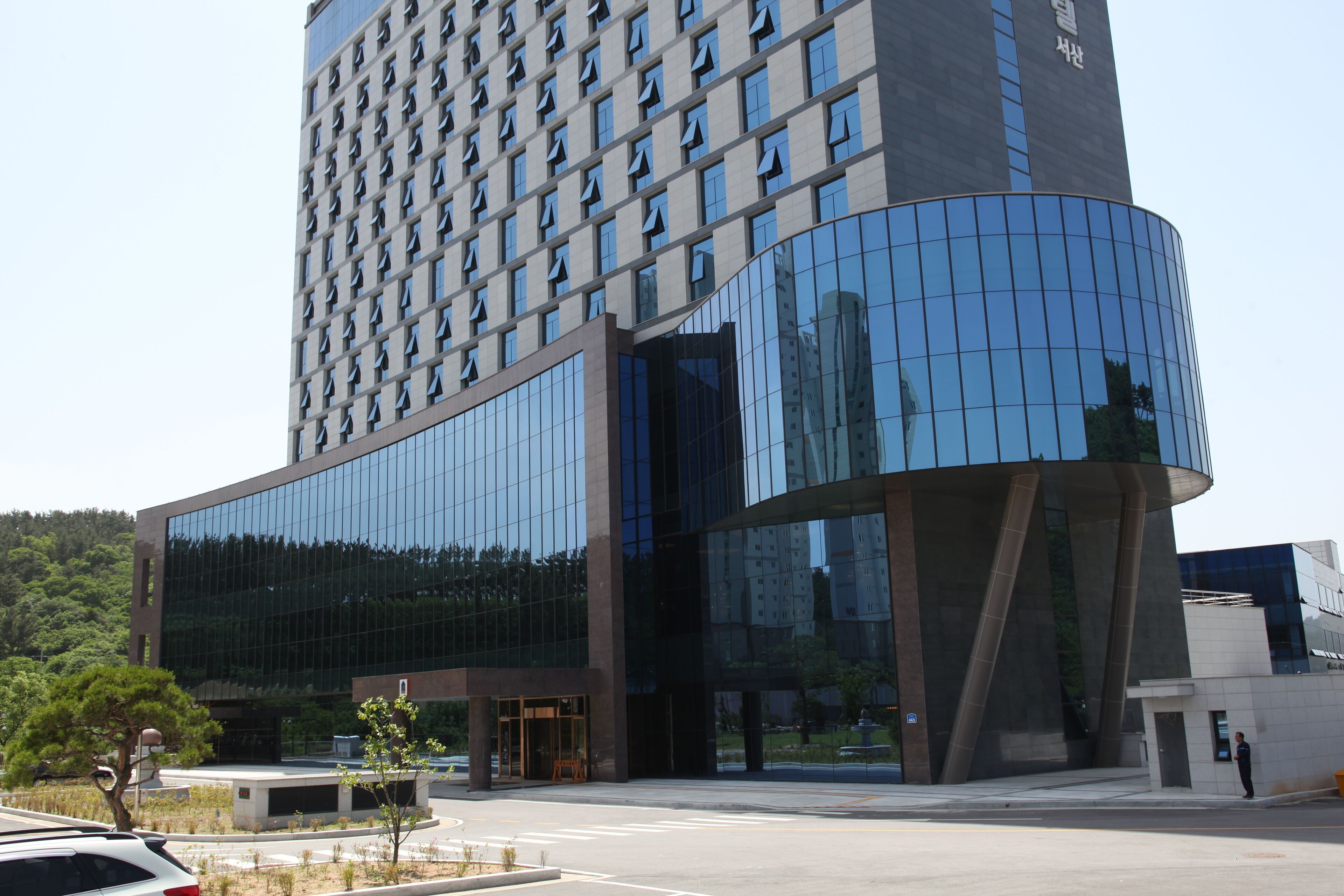 A modern hotel with a curved glass facade, a large entrance, and a parking area with a few cars.