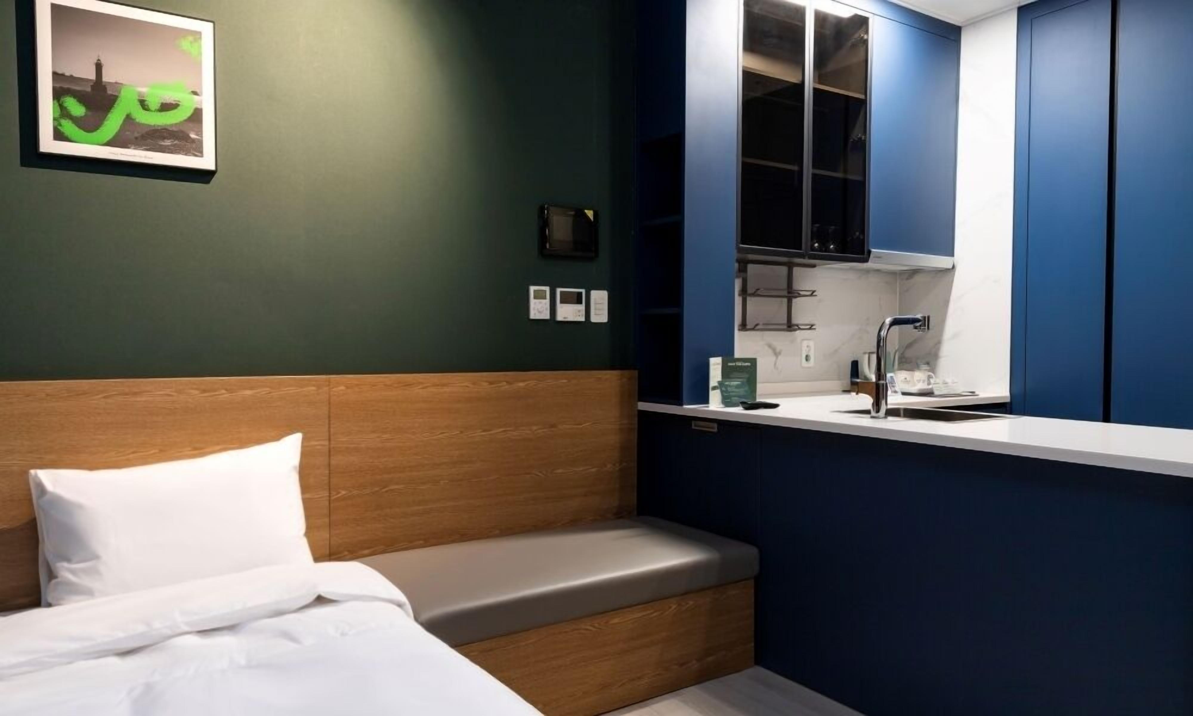 Un dormitorio con cabecera de madera, cama con ropa de cama blanca, un pequeño banco, una ventana con vista a un edificio y un termostato fijado a la pared.