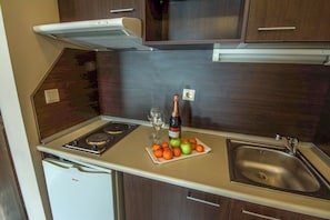 Eine kompakte Küchenzeile mit Herd, Spüle und kleinem Kühlschrank. Auf der Arbeitsfläche steht eine Weinflasche und ein Teller mit Obst.