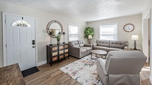 Un salon avec une porte blanche, une table basse en bois, un canapé beige, un canapé gris, une petite table ronde, un miroir, une lampe et une horloge au mur.